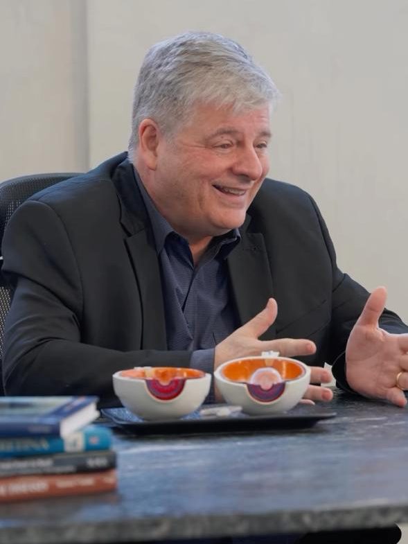 Homem sorridente conversando em uma mesa, com duas tigelas coloridas à frente e livros empilhados ao lado.